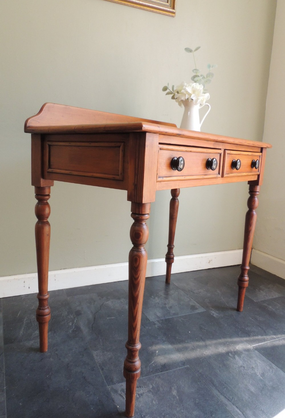 A Victorian solid pine hall table featuring a low gallery backboard, two drawers with dark round knobs, and four turned legs. View XII
