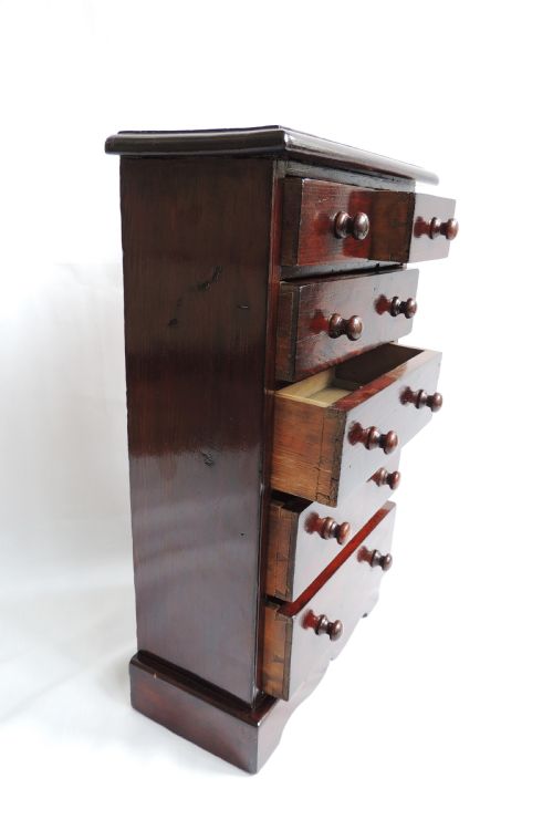 fig 1. A dark stained pine Victorian miniature chest of drawers featuring six drawers with turned wooden knobs and a shaped plinth base.
