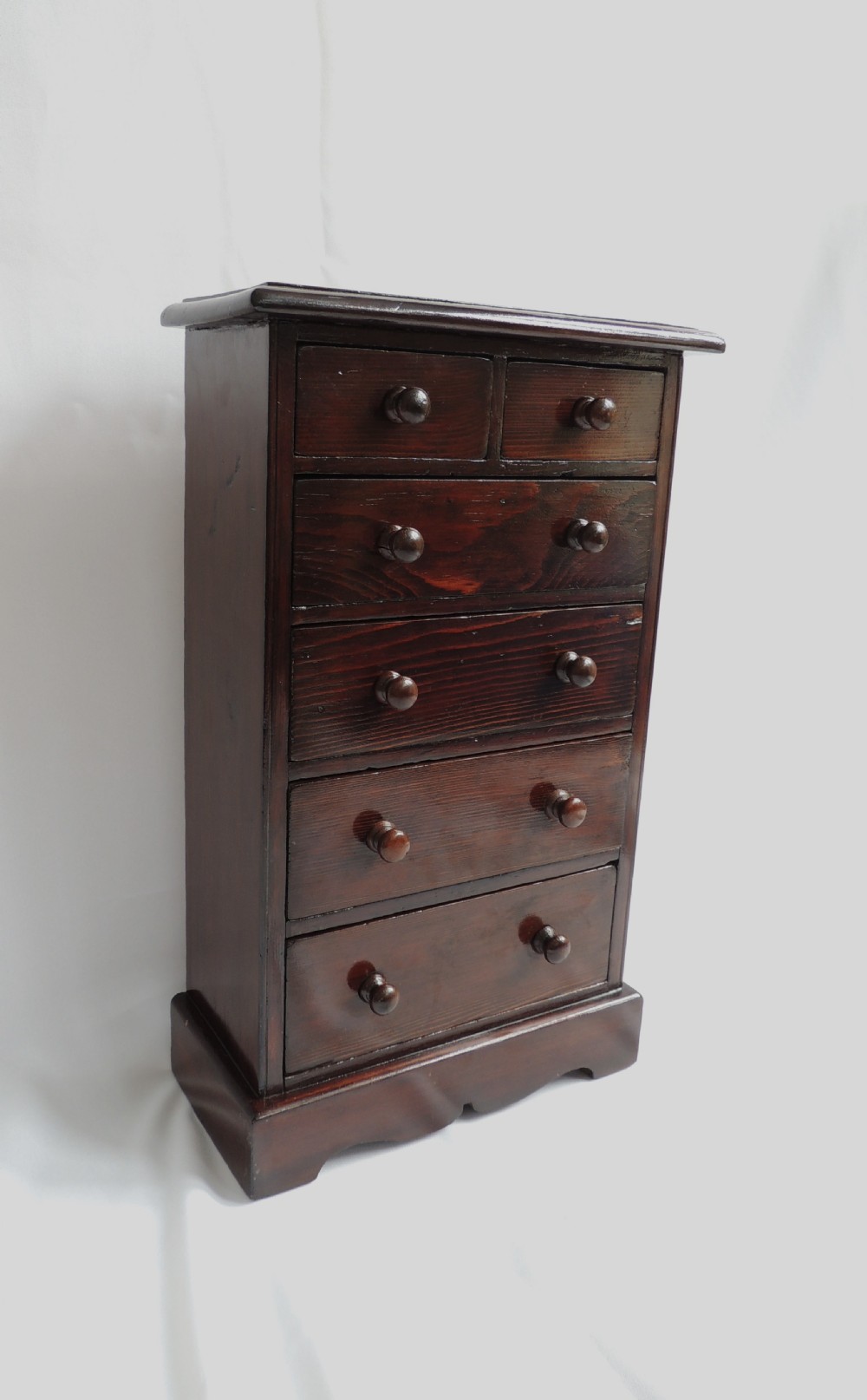 A dark stained pine Victorian miniature chest of drawers featuring six drawers with turned wooden knobs and a shaped plinth base.