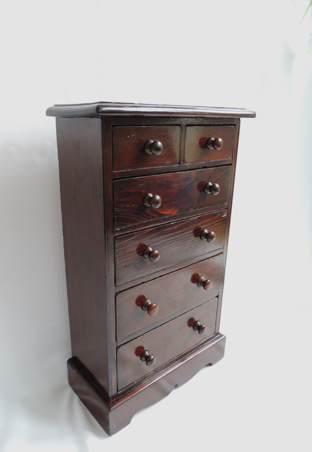 A dark stained pine Victorian miniature chest of drawers featuring six drawers with turned wooden knobs and a shaped plinth base. View IV