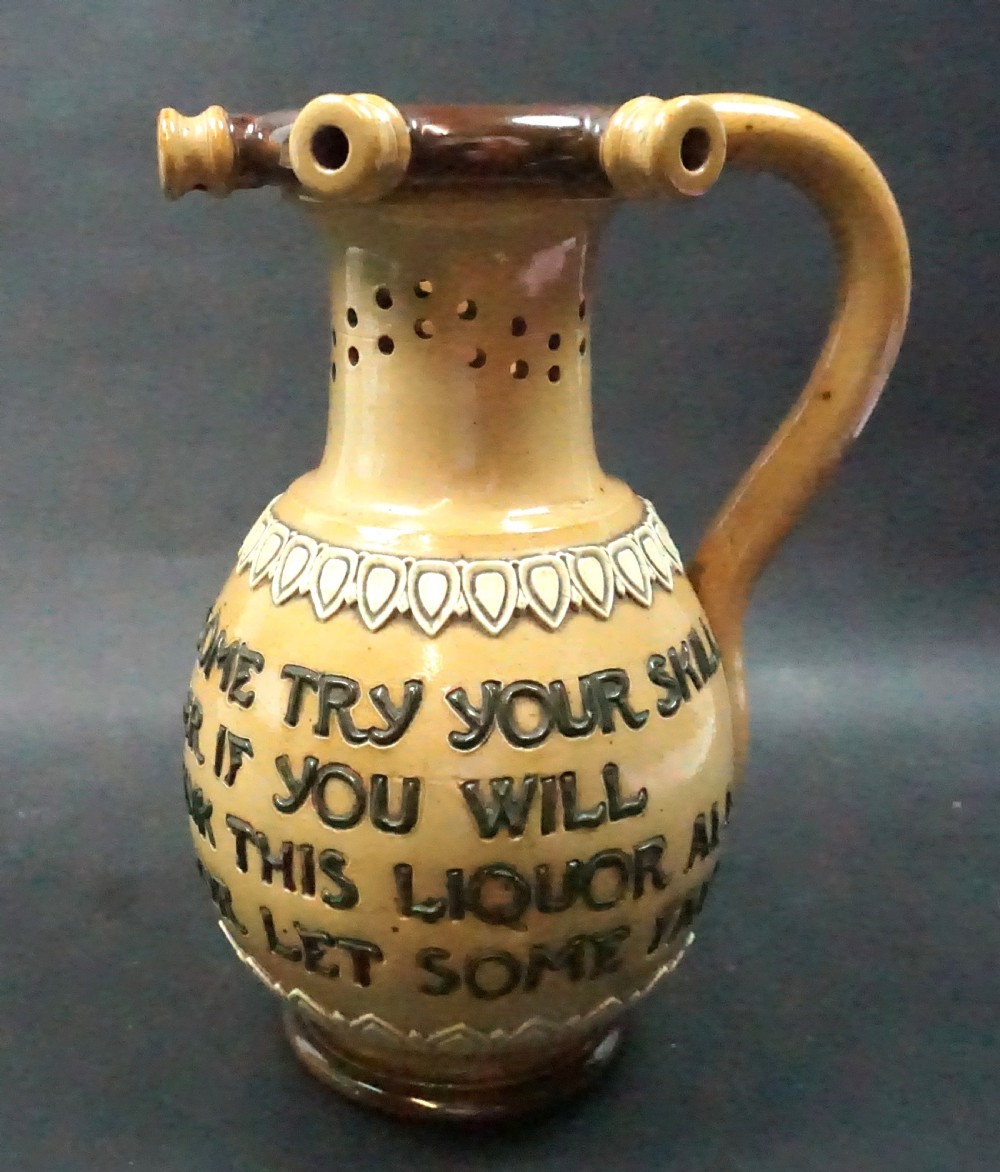 An antique Doulton Lambeth salt glazed stoneware puzzle jug featuring pierced neck detailing, an incised drinking verse on the body, and a dark brown handle.