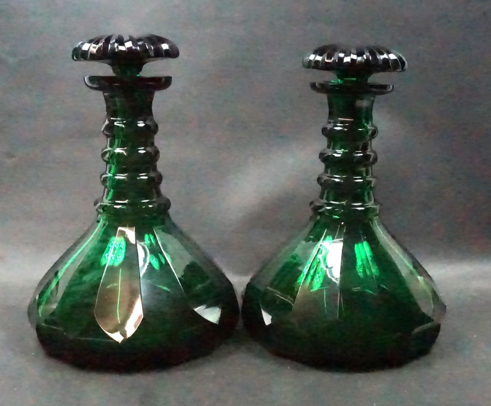 A pair of dark green glass ships decanters featuring broad conical bases with facet cuts, ringed necks, and fitted radial stoppers.