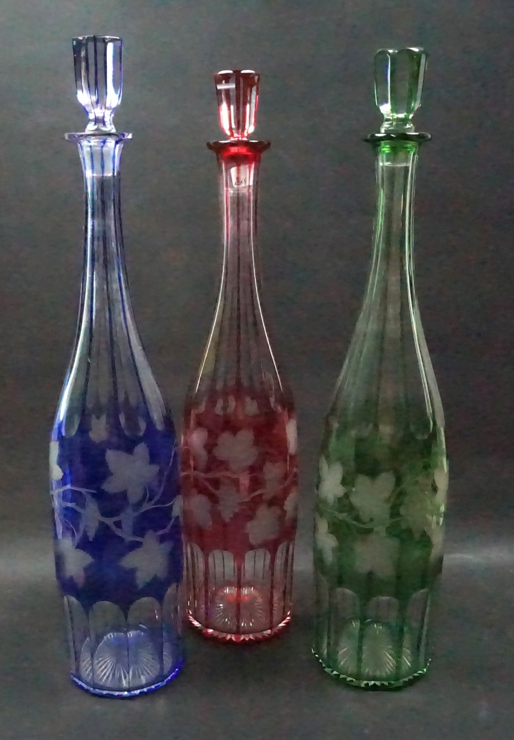 A set of three tall Victorian colour overlay crystal glass hock decanters in blue, ruby red, and emerald green featuring cut vine leaf patterns and faceted stoppers.