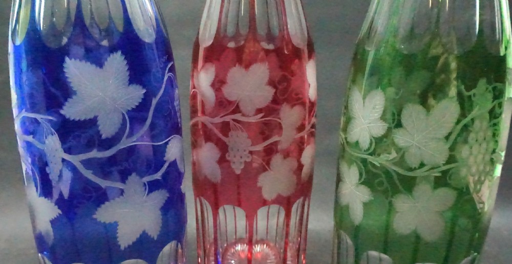 A set of three tall Victorian colour overlay crystal glass hock decanters in blue, ruby red, and emerald green featuring cut vine leaf patterns and faceted stoppers. View II