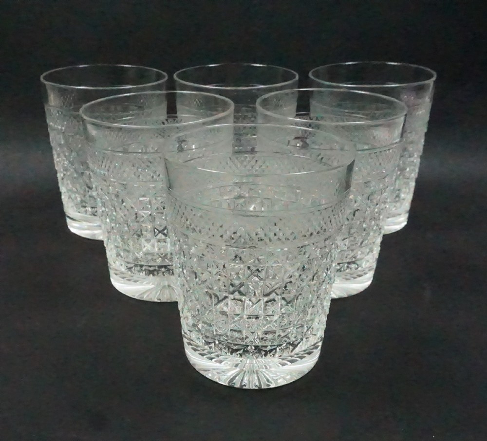 A set of six clear cut crystal whisky tumblers featuring geometric diamond facet cuts and a starburst base pattern arranged on a dark background.