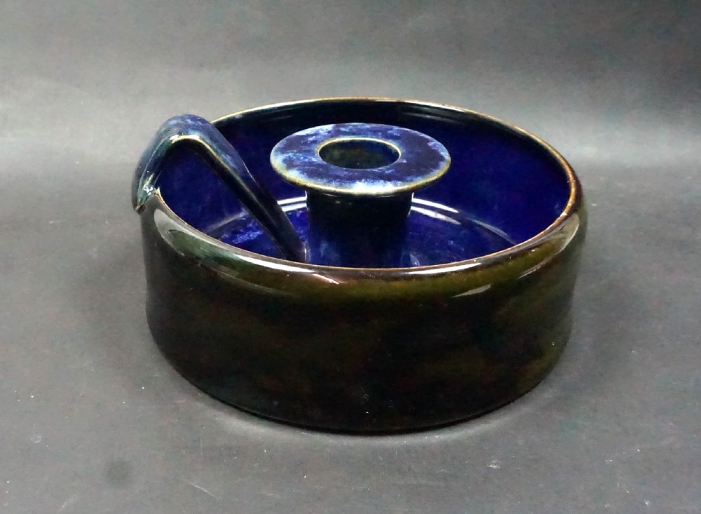 A circular Royal Doulton glazed stoneware chamberstick featuring a central candle sconce, an integrated side ring handle, and a mottled dark blue glaze with olive brown exterior tones.