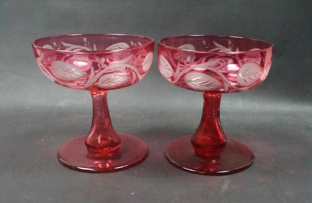 A pair of Victorian cranberry glass champagne coupes featuring hand-engraved convolvulus leaves and vines over clear crystal stems.