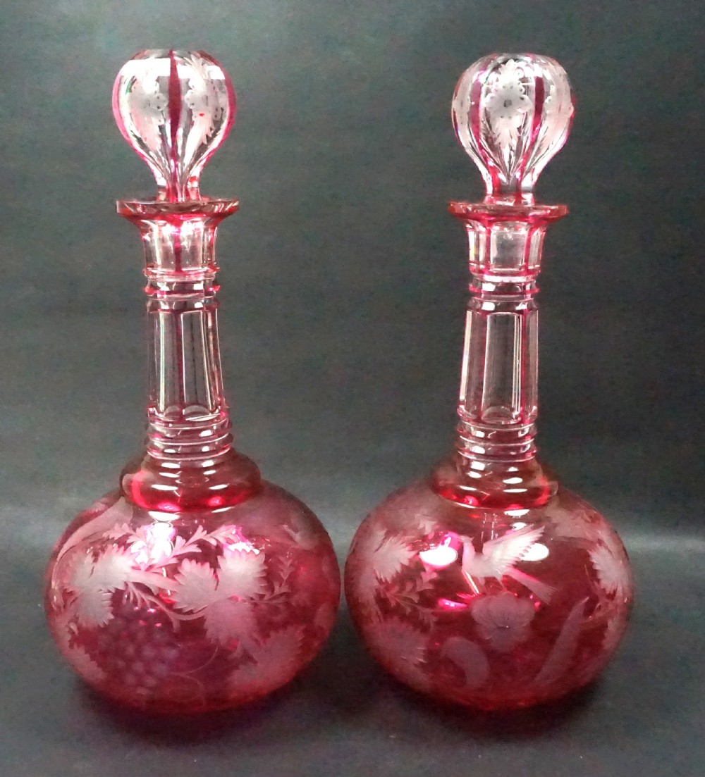 A pair of Victorian cranberry overlay glass decanters featuring cut stoppers and hand-engraved patterns of birds and grapevines.