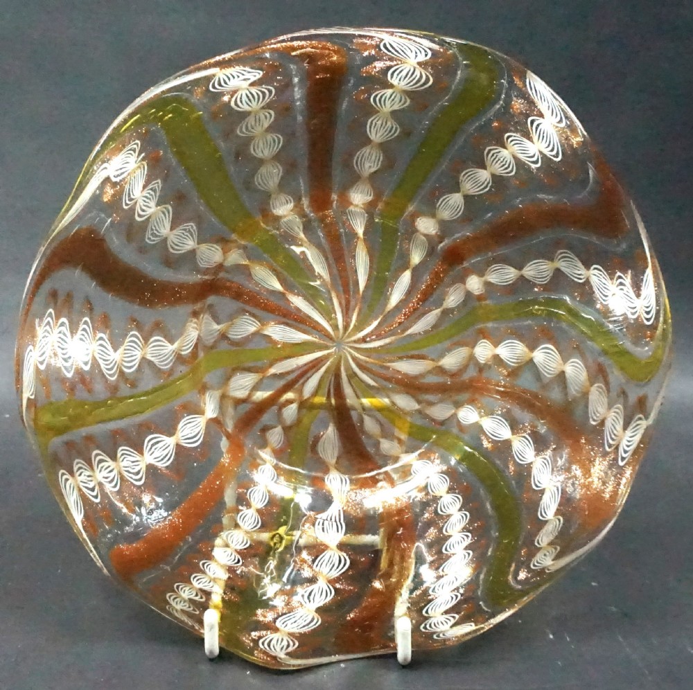 A round Venetian clear glass plate featuring spiral ribbons of green, copper aventurine, and white twisted latticinio patterns expanding from the centre.