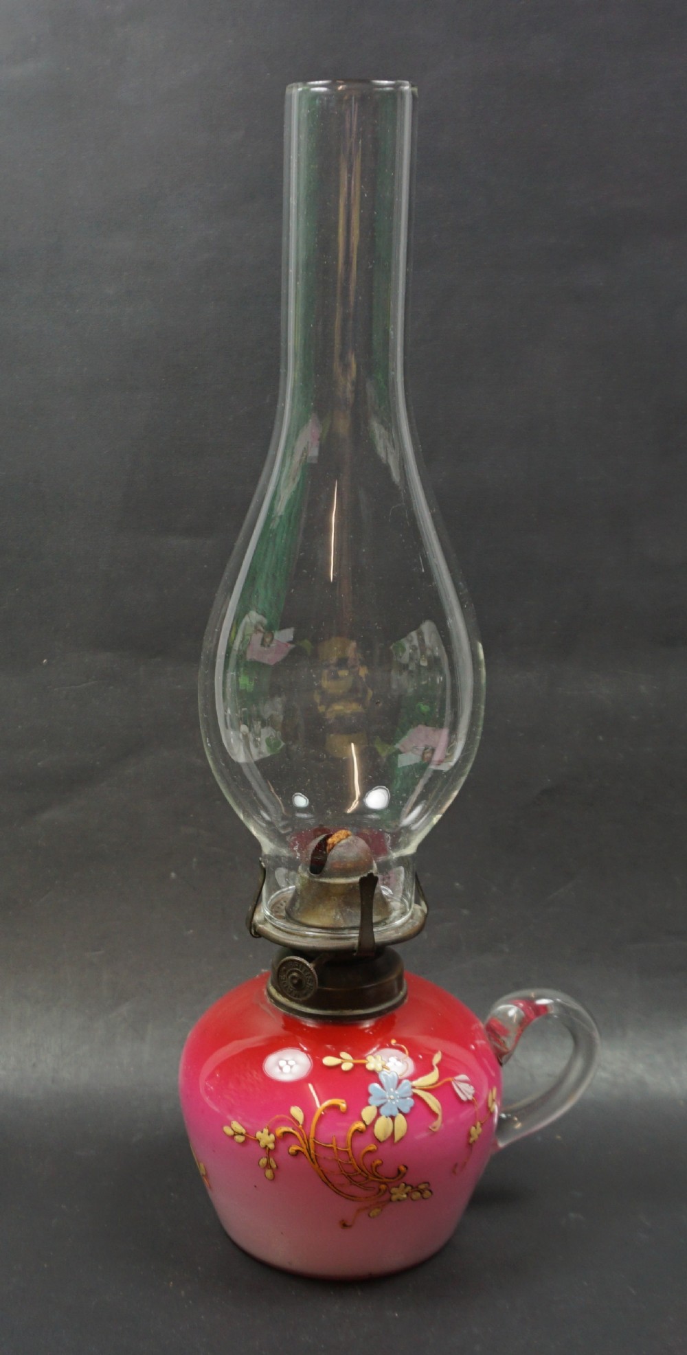 An antique Victorian oil lamp featuring a pink glass reservoir decorated with gilded scrollwork and painted flowers, a clear glass handle, a brass burner, and a tall clear glass chimney.