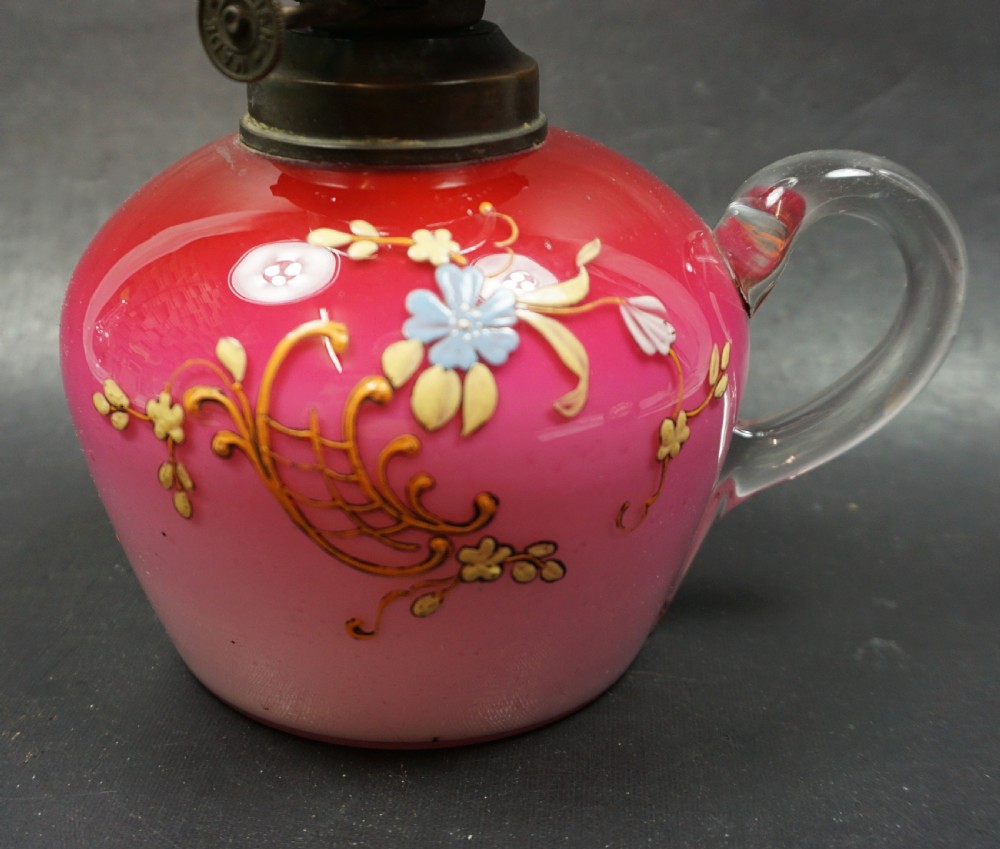 An antique Victorian oil lamp featuring a pink glass reservoir decorated with gilded scrollwork and painted flowers, a clear glass handle, a brass burner, and a tall clear glass chimney. View II