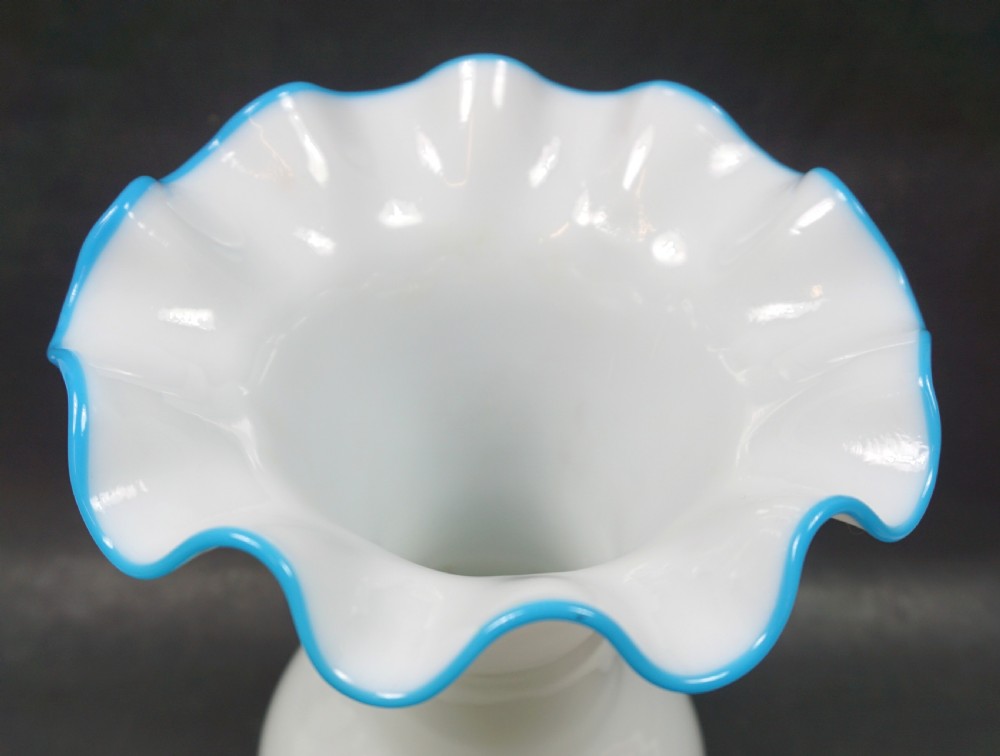 A white opaline glass vase featuring a bulbous base, flared neck, and a ruffled pale blue glass rim. View II