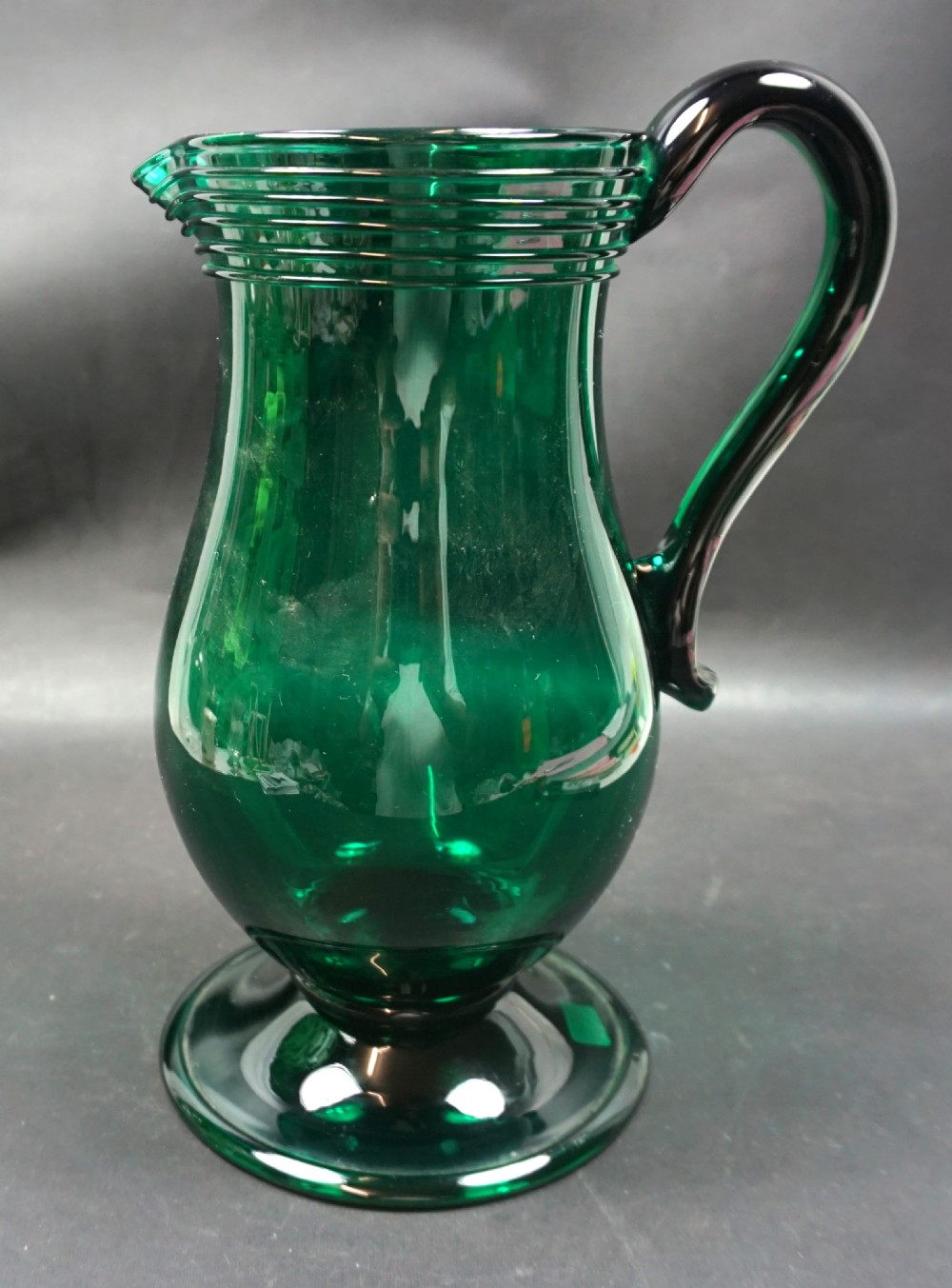 A deep green hand-blown glass jug featuring a folded foot base, a trailed neck rim, and an applied dark handle.