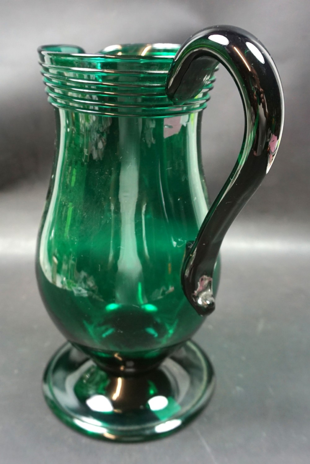 A deep green hand-blown glass jug featuring a folded foot base, a trailed neck rim, and an applied dark handle. View II