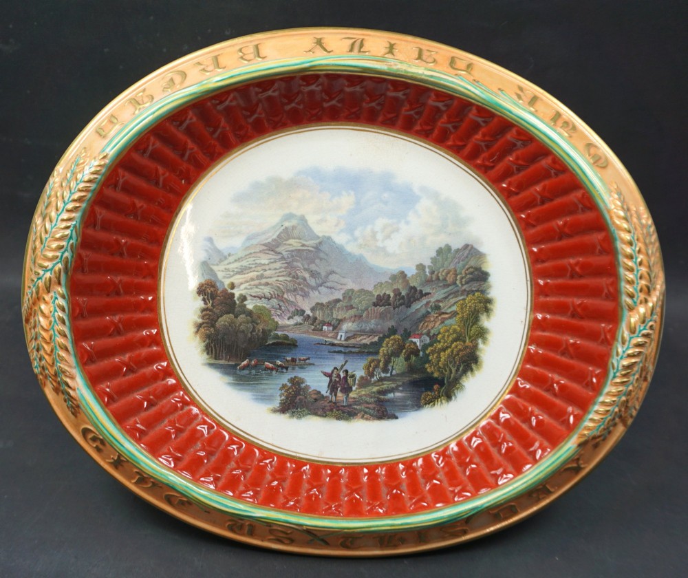 An oval Victorian ceramic bread platter featuring an embossed ochre rim with wheat motif, a red fluted inner border, and a central polychrome transfer landscape of a mountain stream.