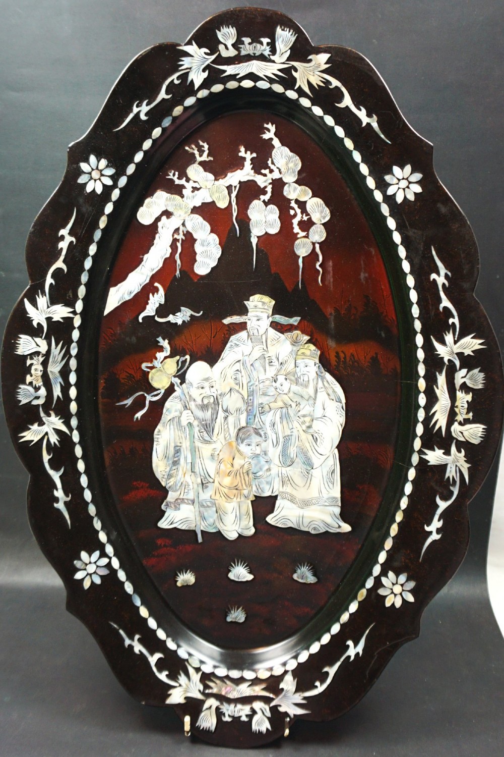 An oval dark red and black lacquered wood platter featuring a scalloped rim and mother of pearl shell inlay depicting figures under a tree.