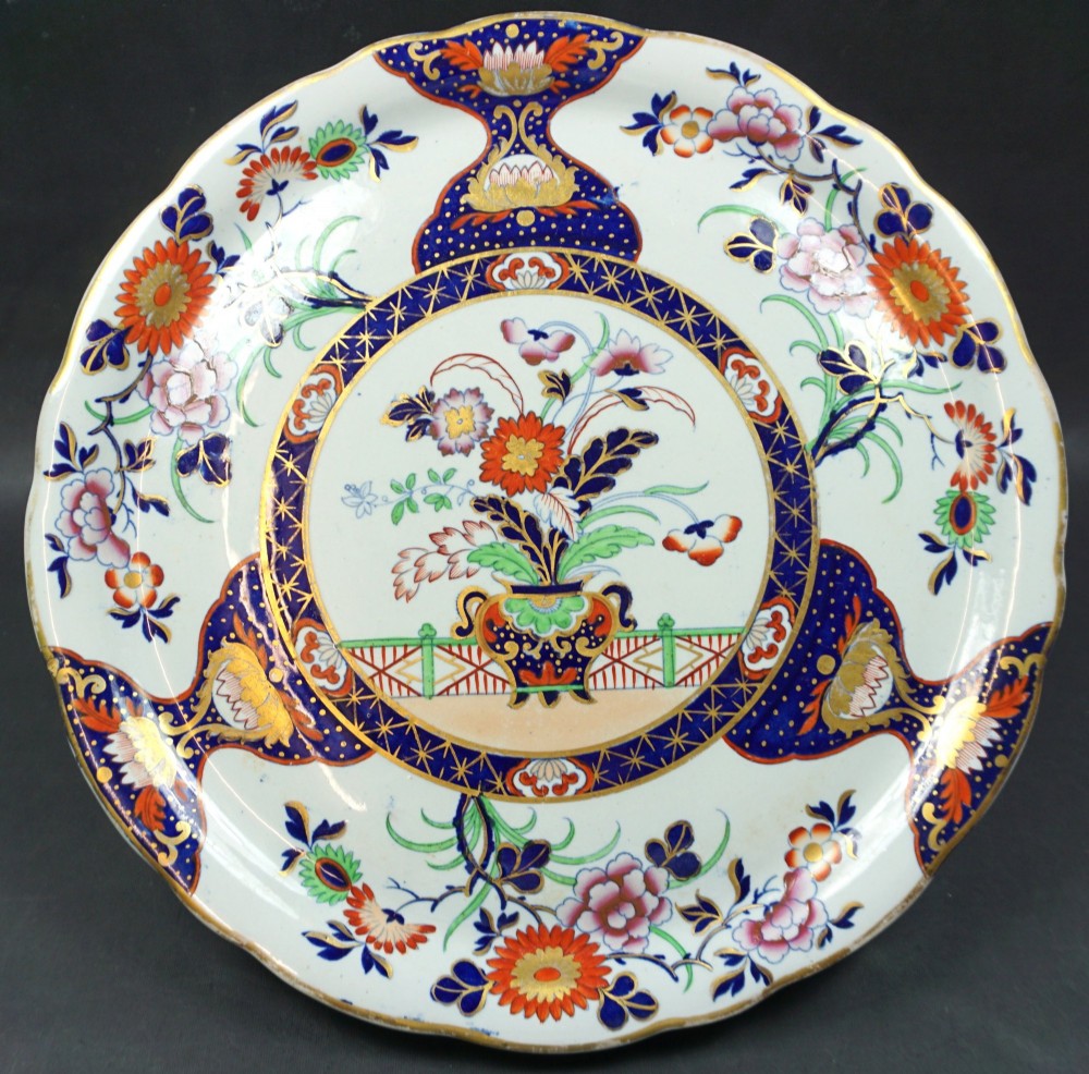 A circular ceramic plate featuring hand-coloured floral Imari patterns in cobalt blue, orange, and pink with a scalloped gilded rim.