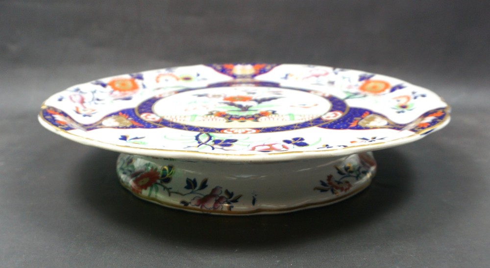 A circular ceramic plate featuring hand-coloured floral Imari patterns in cobalt blue, orange, and pink with a scalloped gilded rim. View II