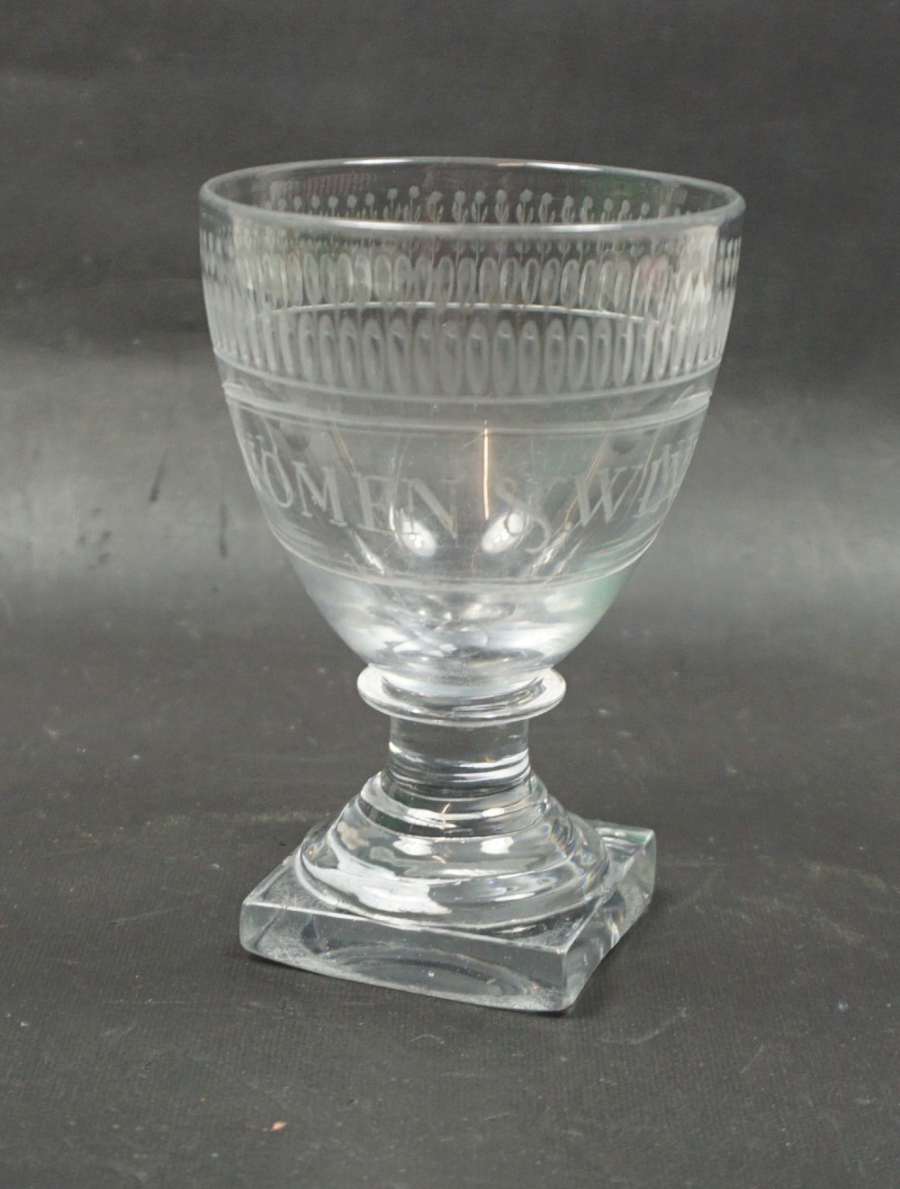 A clear Georgian glass rummer featuring a square foot, turned stem, and an engraved bowl inscribed with the words Women and Wine beneath a decorative rim band.