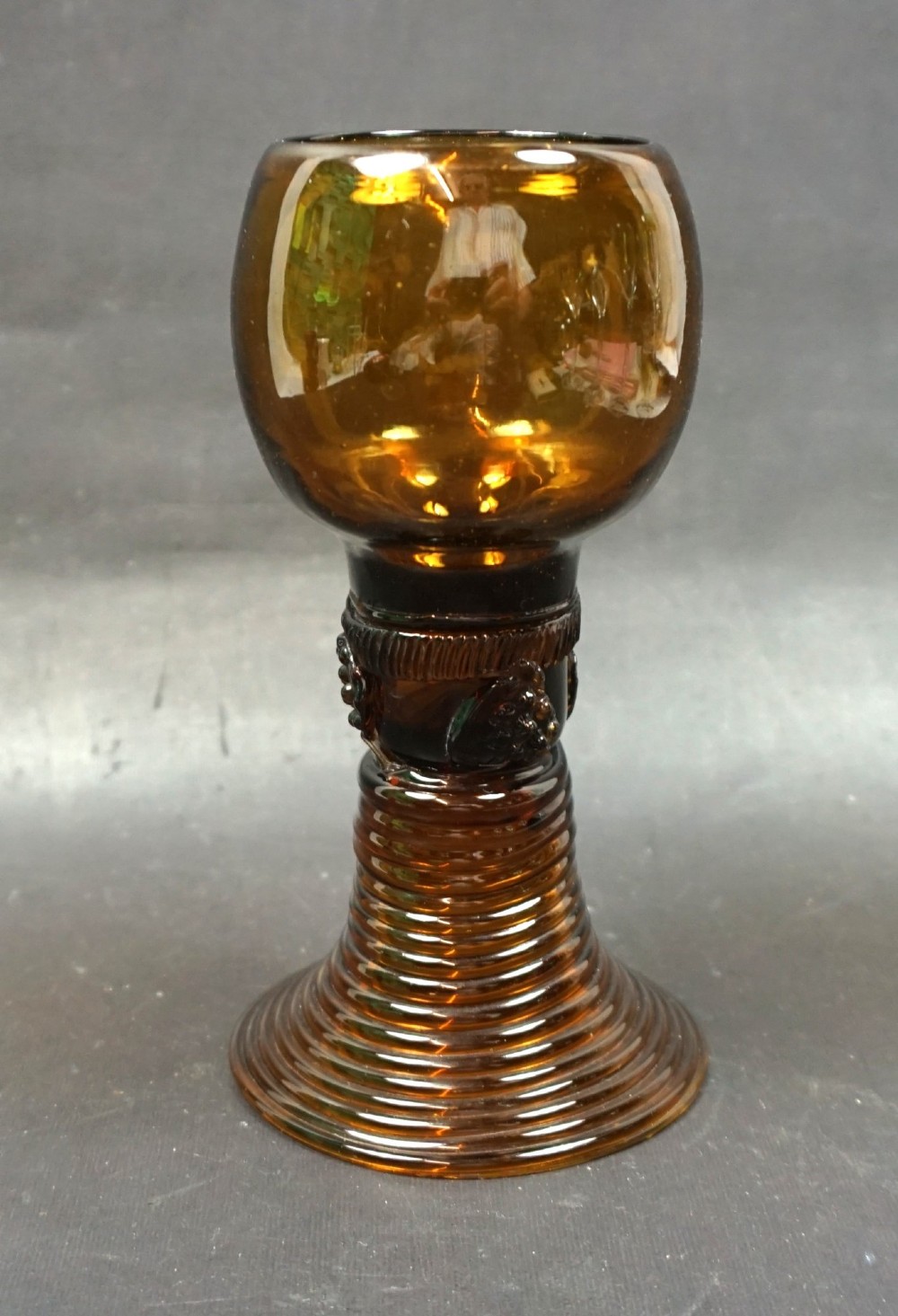 A dark amber glass roemer featuring a rounded bowl, a prunted stem, and a ribbed coiled glass foot.