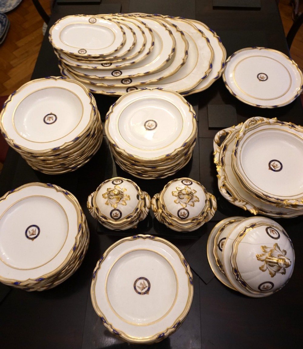 A comprehensive Victorian Staffordshire porcelain dinner service arranged on a dark surface featuring white plates, serving platters, and covered tureens decorated with cobalt blue trim and gilded heraldic crests.
