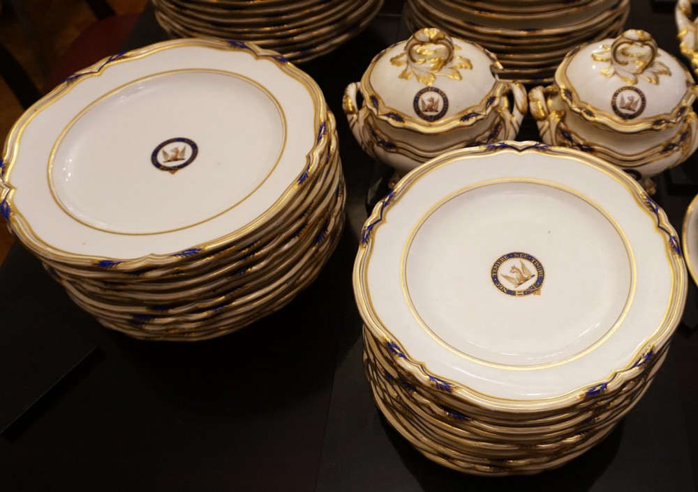 A comprehensive Victorian Staffordshire porcelain dinner service arranged on a dark surface featuring white plates, serving platters, and covered tureens decorated with cobalt blue trim and gilded heraldic crests. View II