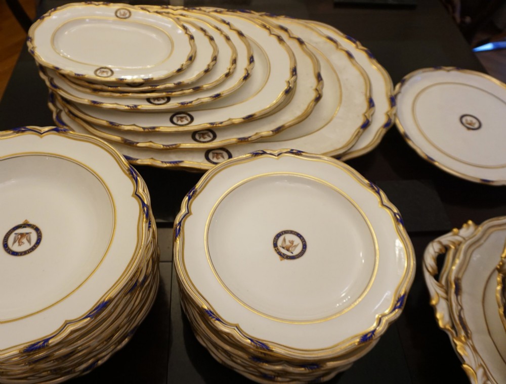 A comprehensive Victorian Staffordshire porcelain dinner service arranged on a dark surface featuring white plates, serving platters, and covered tureens decorated with cobalt blue trim and gilded heraldic crests. View III