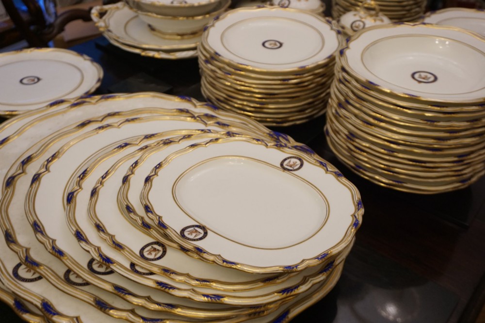 A comprehensive Victorian Staffordshire porcelain dinner service arranged on a dark surface featuring white plates, serving platters, and covered tureens decorated with cobalt blue trim and gilded heraldic crests. View IV