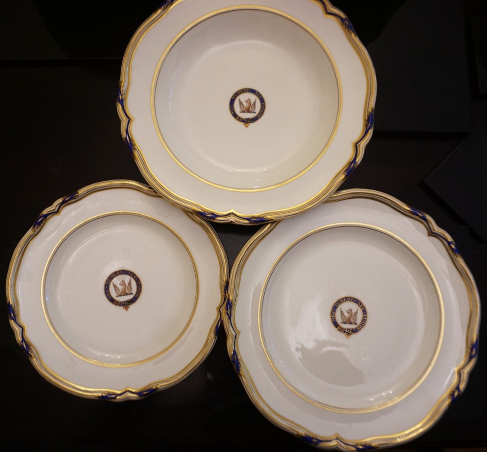 A comprehensive Victorian Staffordshire porcelain dinner service arranged on a dark surface featuring white plates, serving platters, and covered tureens decorated with cobalt blue trim and gilded heraldic crests. View VI