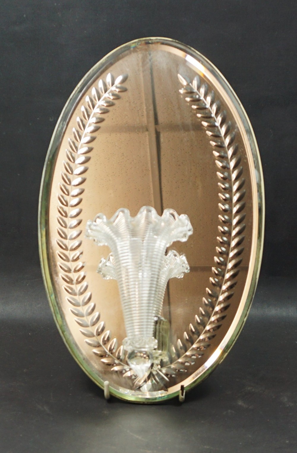 An oval silvered glass wall plaque featuring engraved leaf borders and a centrally mounted ribbed glass posey vase with a fluted rim.