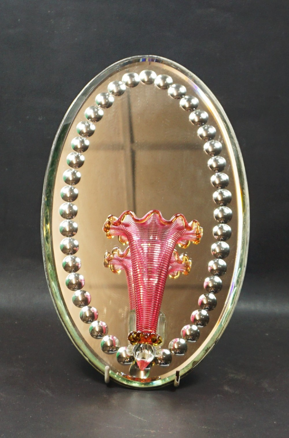An oval mirrored wall plaque bordered by silvered glass cabochons featuring an attached central pink cranberry glass trumpet vase with a crimped rim.