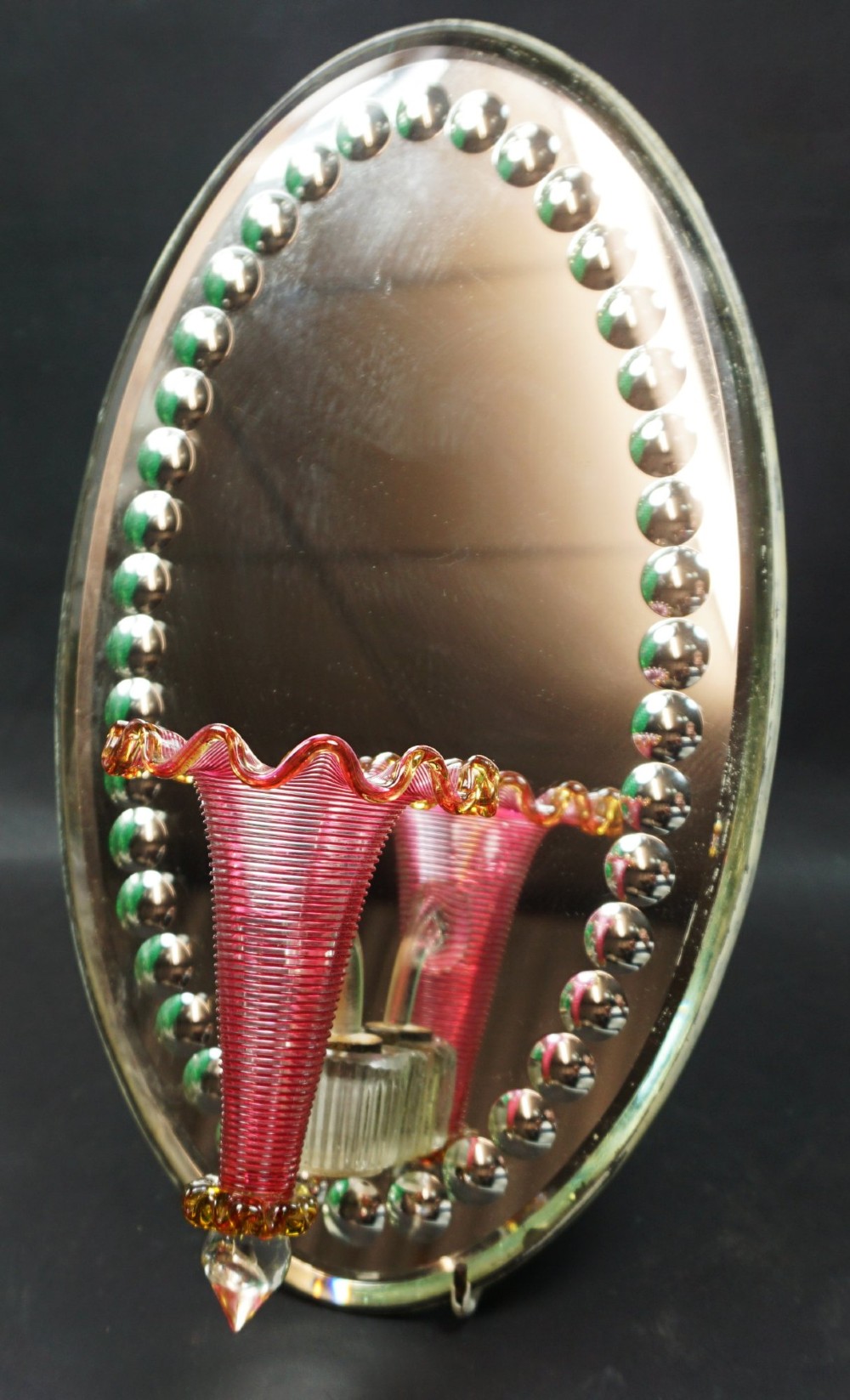 An oval mirrored wall plaque bordered by silvered glass cabochons featuring an attached central pink cranberry glass trumpet vase with a crimped rim. View II