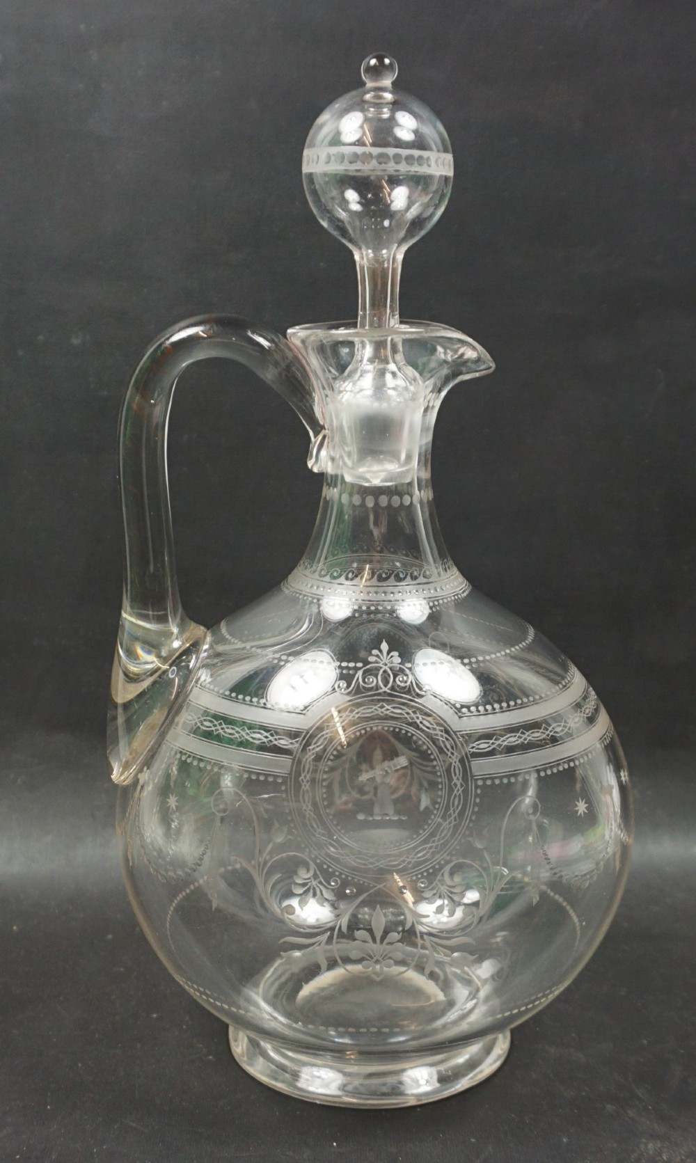 An antique clear glass wine decanter featuring wheel-engraved heraldic crests, a rounded loop handle, and a matching spherical stopper.