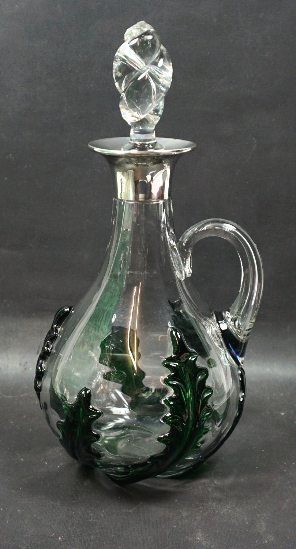 A clear crystal glass decanter featuring dark green applied leaf mouldings around the lower body, a loop handle, a silver collar, and a twisted cut-glass stopper.