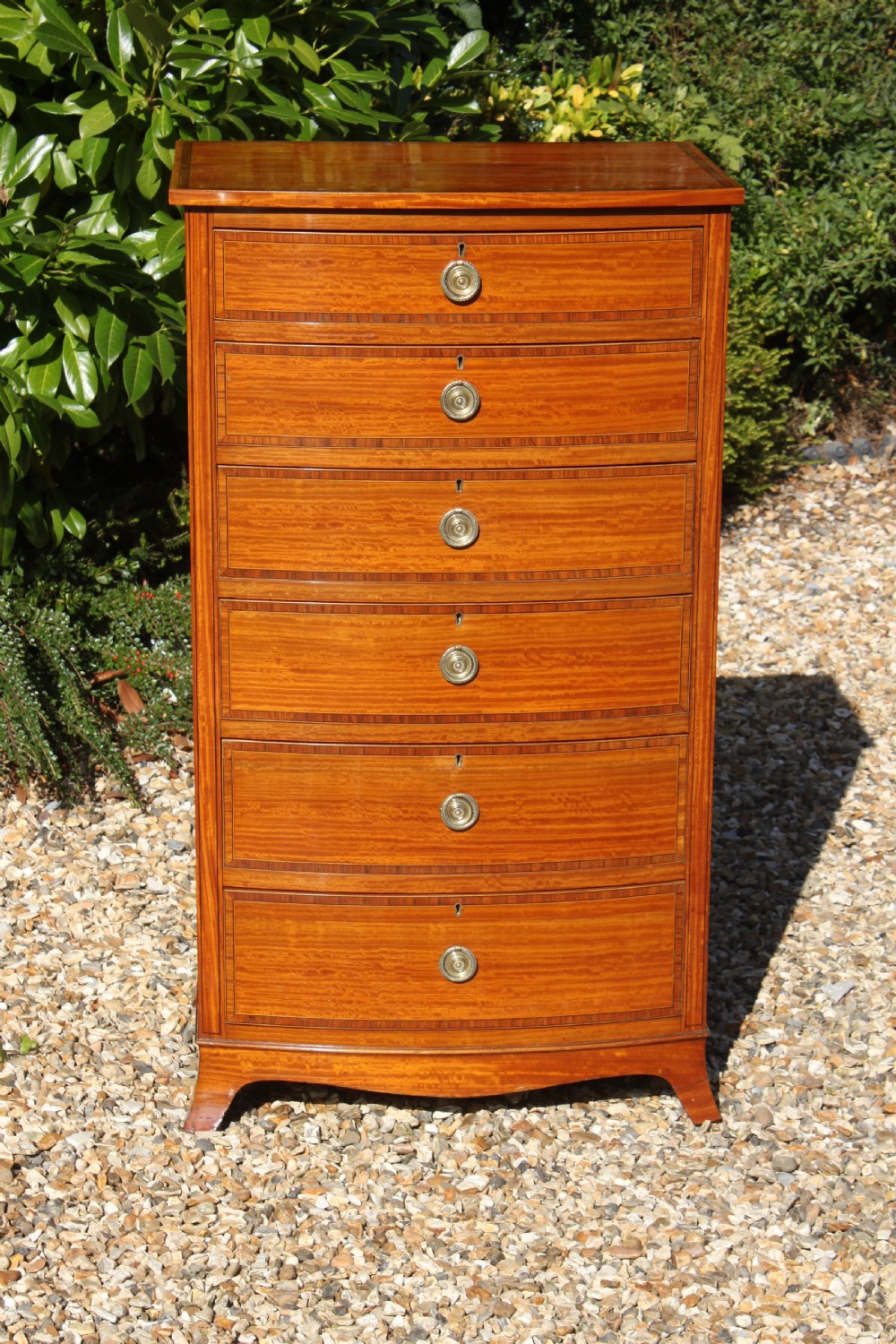 A tall Sheraton Revival satinwood bow front chest featuring six drawers with inlaid crossbanded borders, central brass ring handles, and splayed bracket feet.
