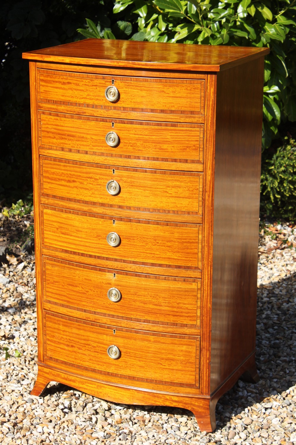 A tall Sheraton Revival satinwood bow front chest featuring six drawers with inlaid crossbanded borders, central brass ring handles, and splayed bracket feet. View V