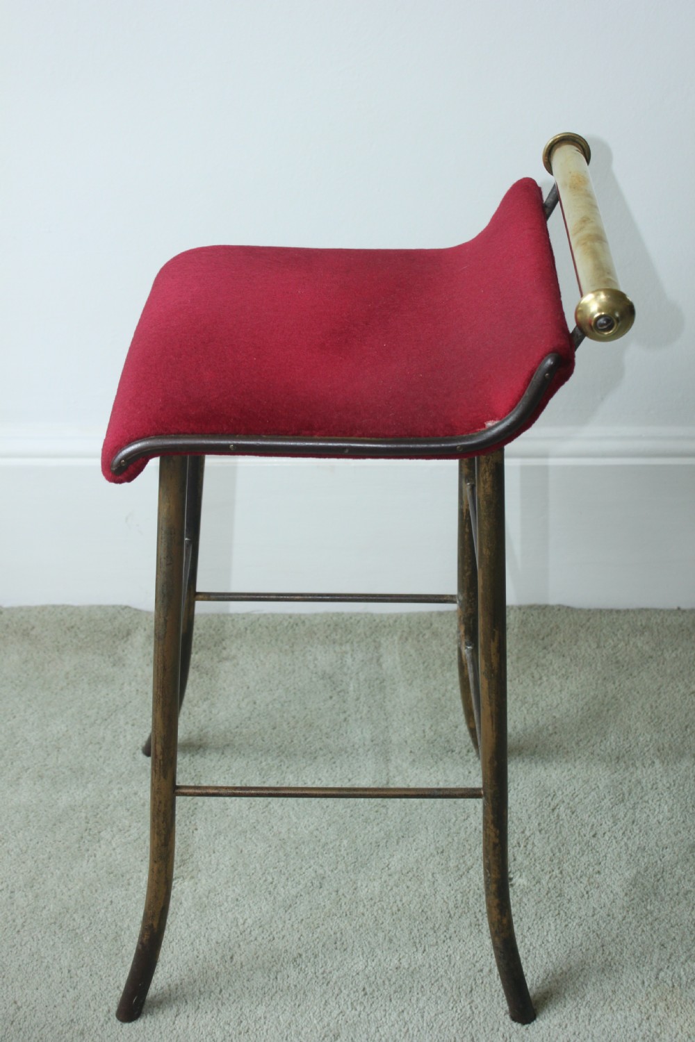 A 19th-century metal music stool featuring a red velvet seat, curved steel legs, and a cylindrical brass top rail with end finials. View V