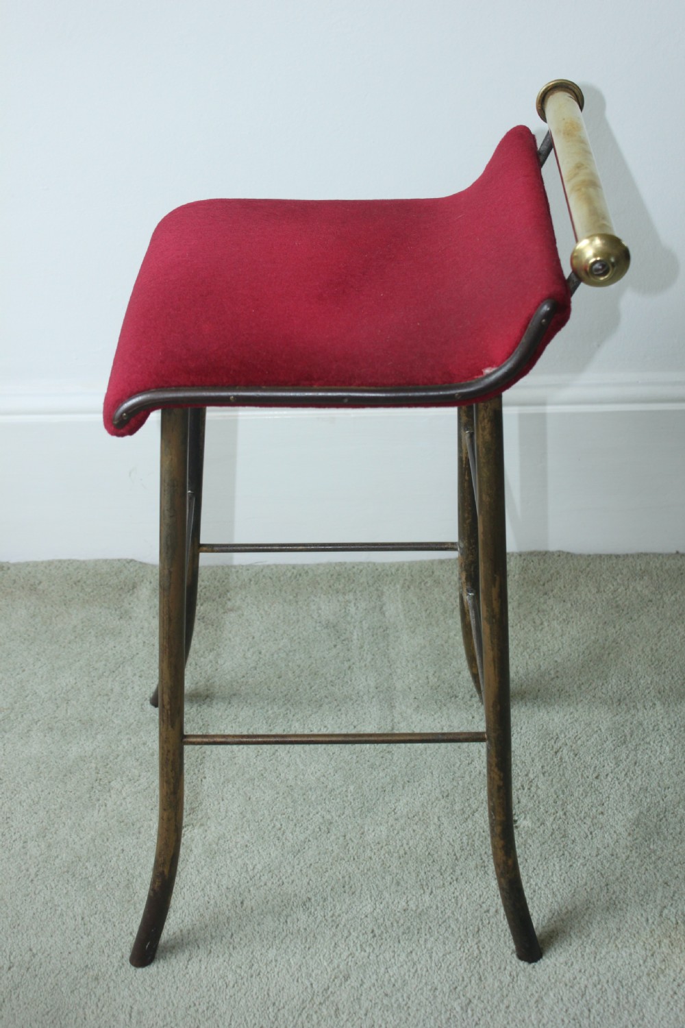 A 19th-century metal music stool featuring a red velvet seat, curved steel legs, and a cylindrical brass top rail with end finials. View VI