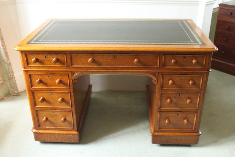 A 19th-century oak pedestal desk featuring a dark green leather writing surface with gold tool gilding, nine drawers with turned wooden knobs, and decorative carved kneehole spandrels.