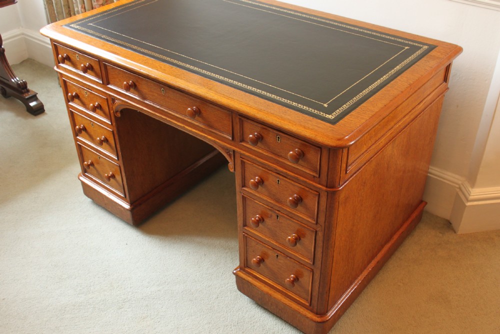 A 19th-century oak pedestal desk featuring a dark green leather writing surface with gold tool gilding, nine drawers with turned wooden knobs, and decorative carved kneehole spandrels. View III