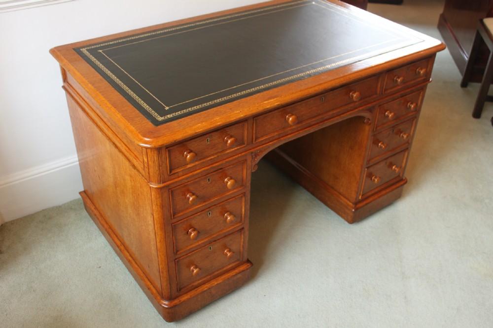 A 19th-century oak pedestal desk featuring a dark green leather writing surface with gold tool gilding, nine drawers with turned wooden knobs, and decorative carved kneehole spandrels. View V