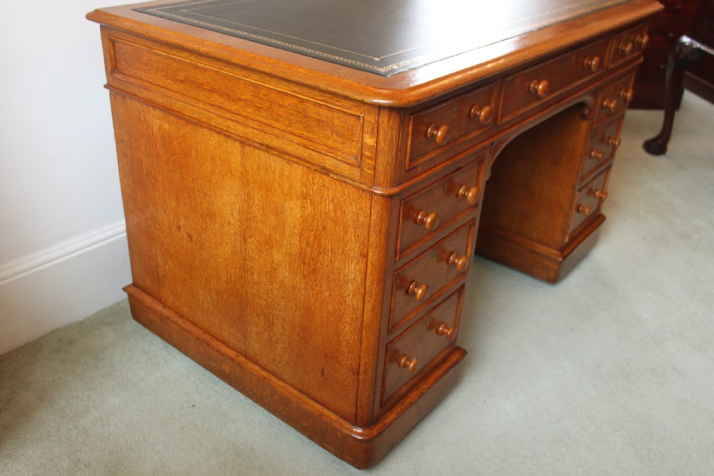 A 19th-century oak pedestal desk featuring a dark green leather writing surface with gold tool gilding, nine drawers with turned wooden knobs, and decorative carved kneehole spandrels. View VI