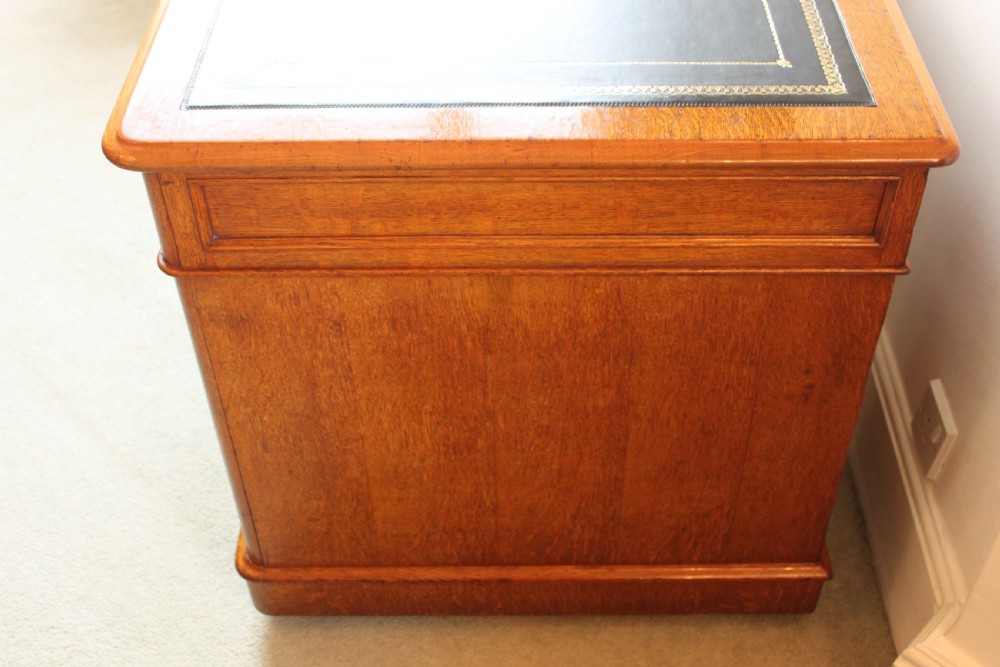 A 19th-century oak pedestal desk featuring a dark green leather writing surface with gold tool gilding, nine drawers with turned wooden knobs, and decorative carved kneehole spandrels. View VII