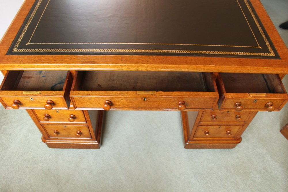 A 19th-century oak pedestal desk featuring a dark green leather writing surface with gold tool gilding, nine drawers with turned wooden knobs, and decorative carved kneehole spandrels. View XI