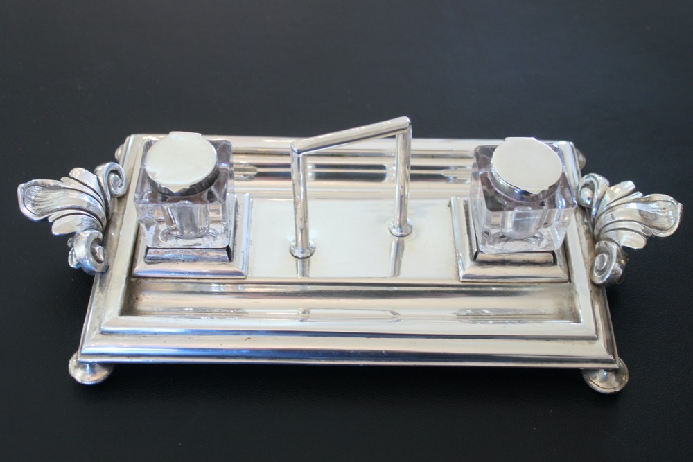 An antique silver-plated rectangular inkstand featuring two square glass inkwells with hinged metal lids, a central loop handle, and decorative side handles.