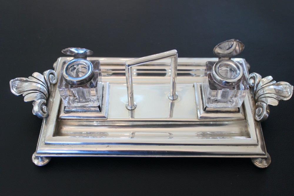 An antique silver-plated rectangular inkstand featuring two square glass inkwells with hinged metal lids, a central loop handle, and decorative side handles. View II