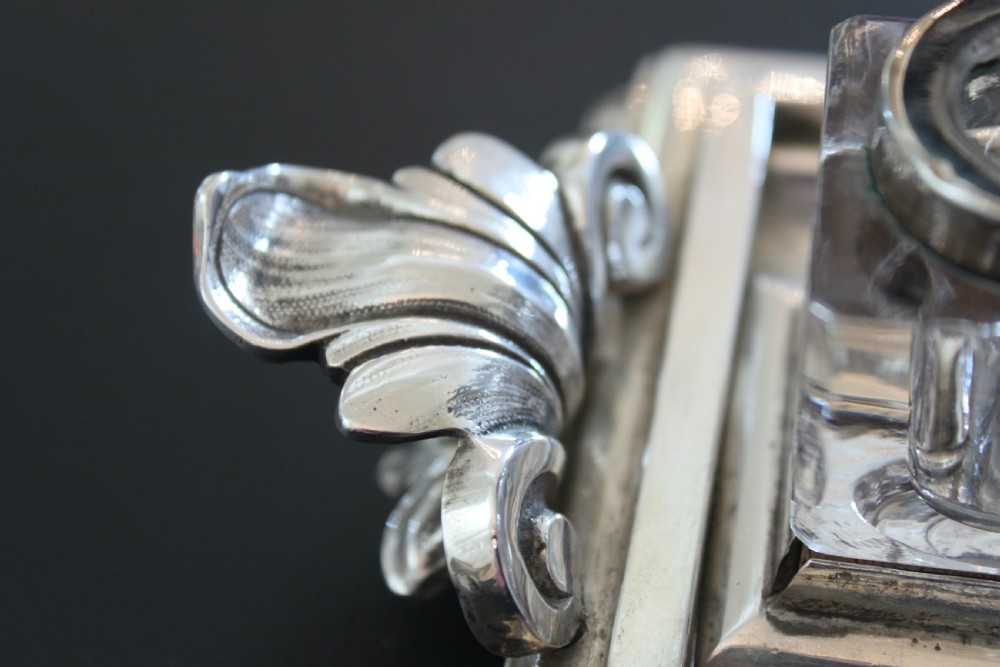 An antique silver-plated rectangular inkstand featuring two square glass inkwells with hinged metal lids, a central loop handle, and decorative side handles. View III