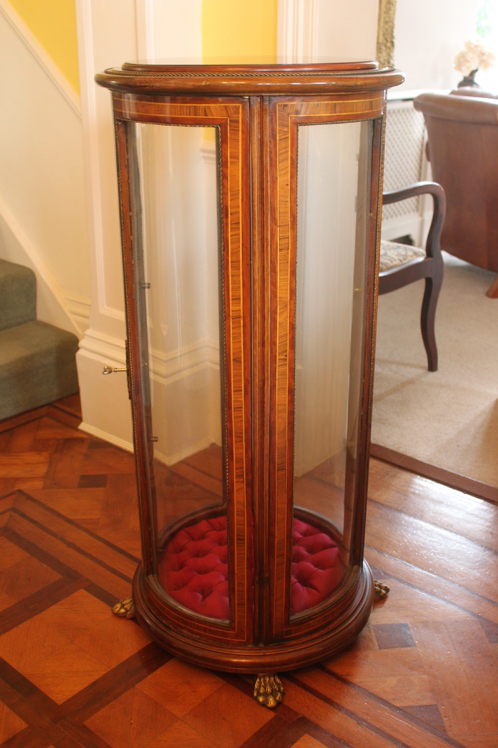 A late 19th-century cylindrical French vitrine cabinet featuring inlaid timber patterning, curved glass panels, tufted red velvet interior lining, and brass claw feet.