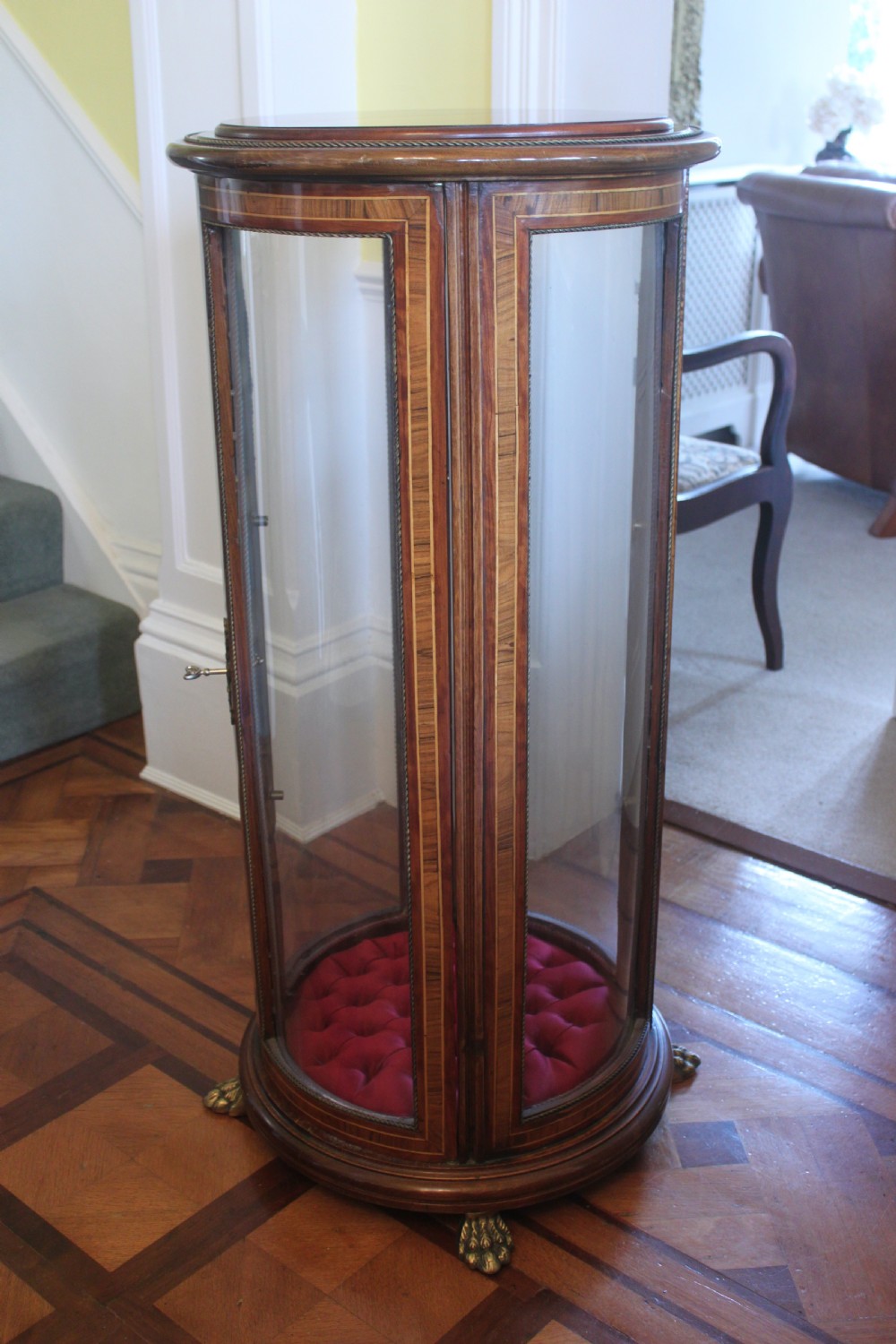 A late 19th-century cylindrical French vitrine cabinet featuring inlaid timber patterning, curved glass panels, tufted red velvet interior lining, and brass claw feet. View II