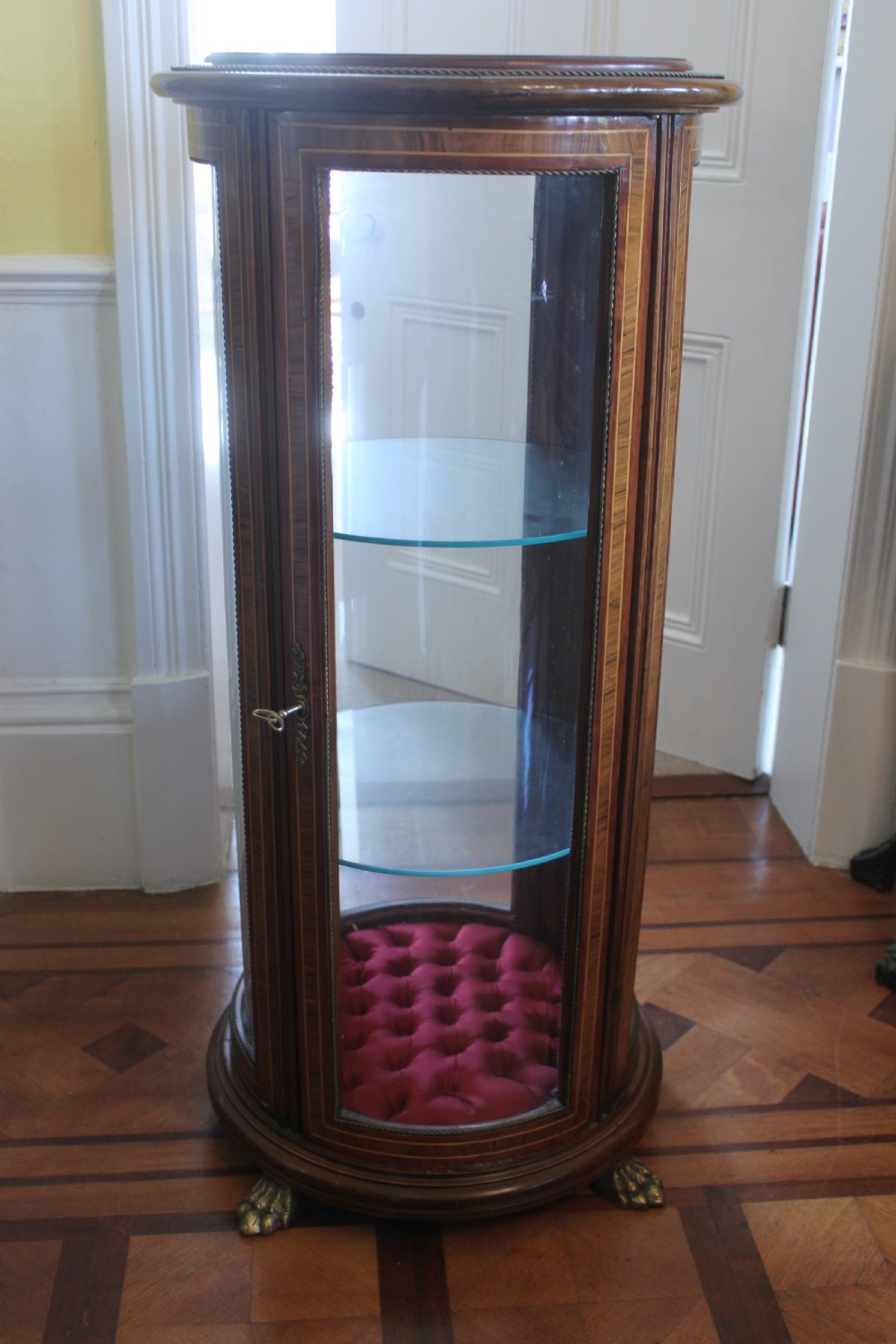 A late 19th-century cylindrical French vitrine cabinet featuring inlaid timber patterning, curved glass panels, tufted red velvet interior lining, and brass claw feet. View III